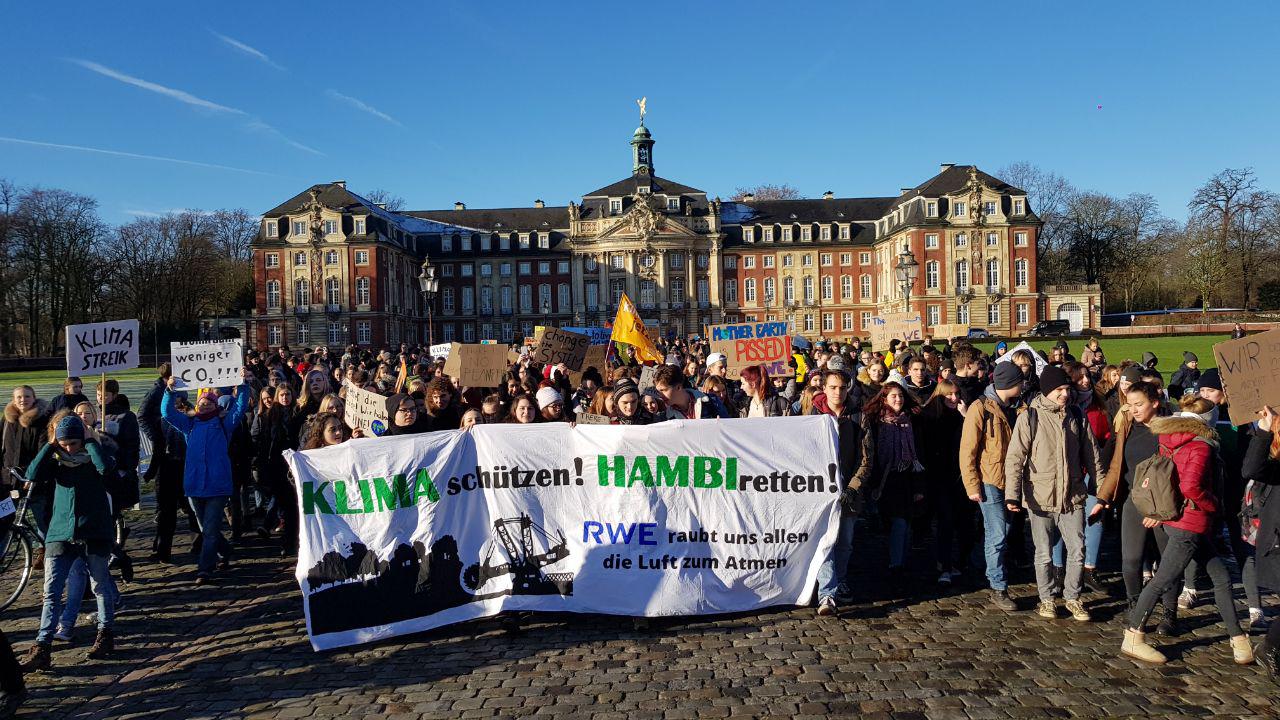 Viele Menschen stehen vor dem Schloss in Münster. Auf einem großen Banner steht: "Klima schützen! Hambi retten! RWE raubt uns allen die Luft zum Atmen"
