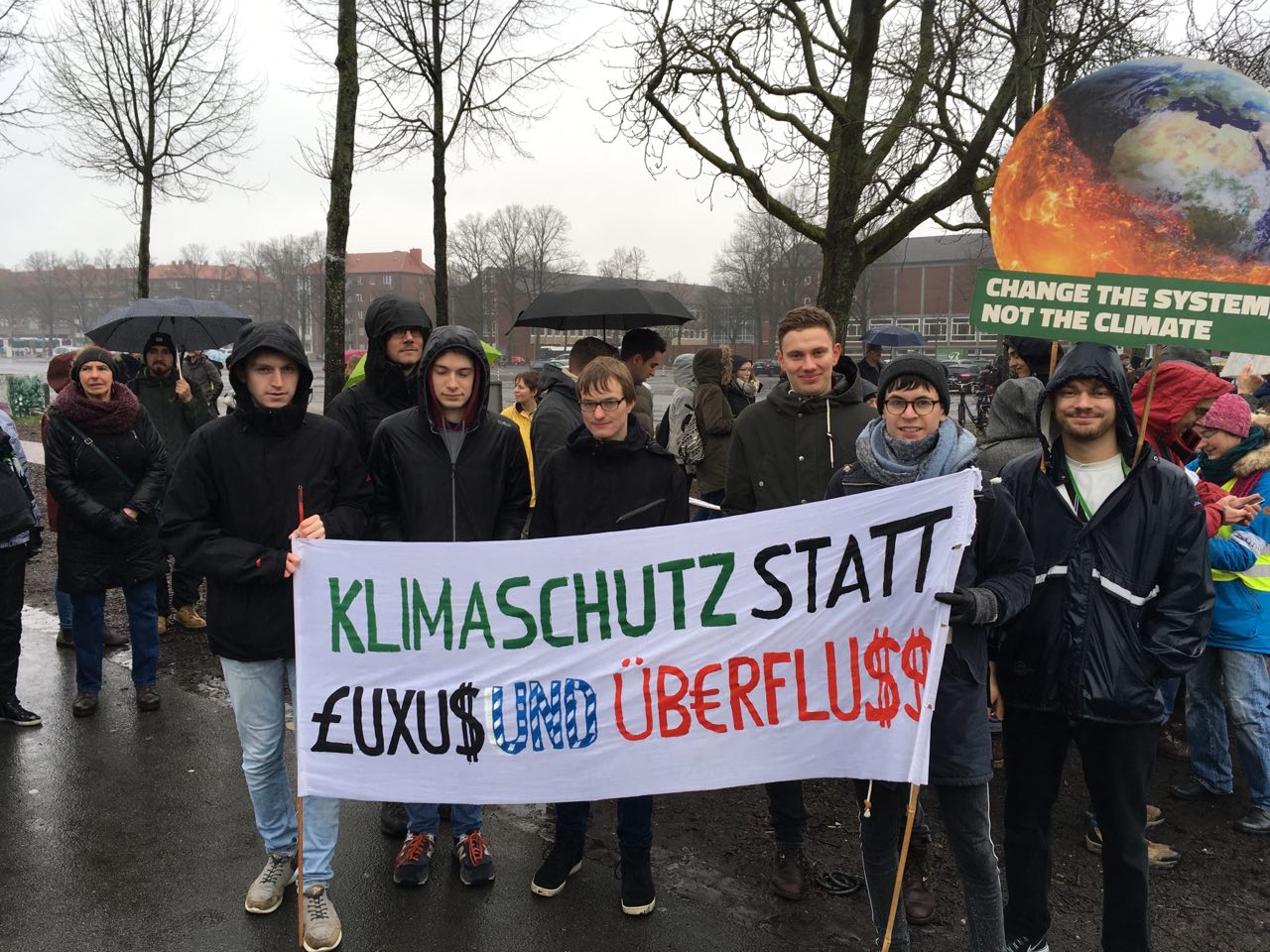 Viele Menschen sind auf einer Demonstration im Regen. Im Vordergrund stehen einige Mitglieder des Kaktus. Sie halten zwei Schilder. Auf einem steht: "Klimaschutz statt Luxus und Überfluss." Auf dem anderen steht: "Change the system, not the climate"