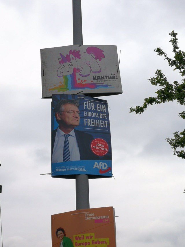 Auf einem Plakat des Kaktus ist ein kotzendes Einhorn. Das Einhorn kotzt auf ein Plakat der AfD herunter.