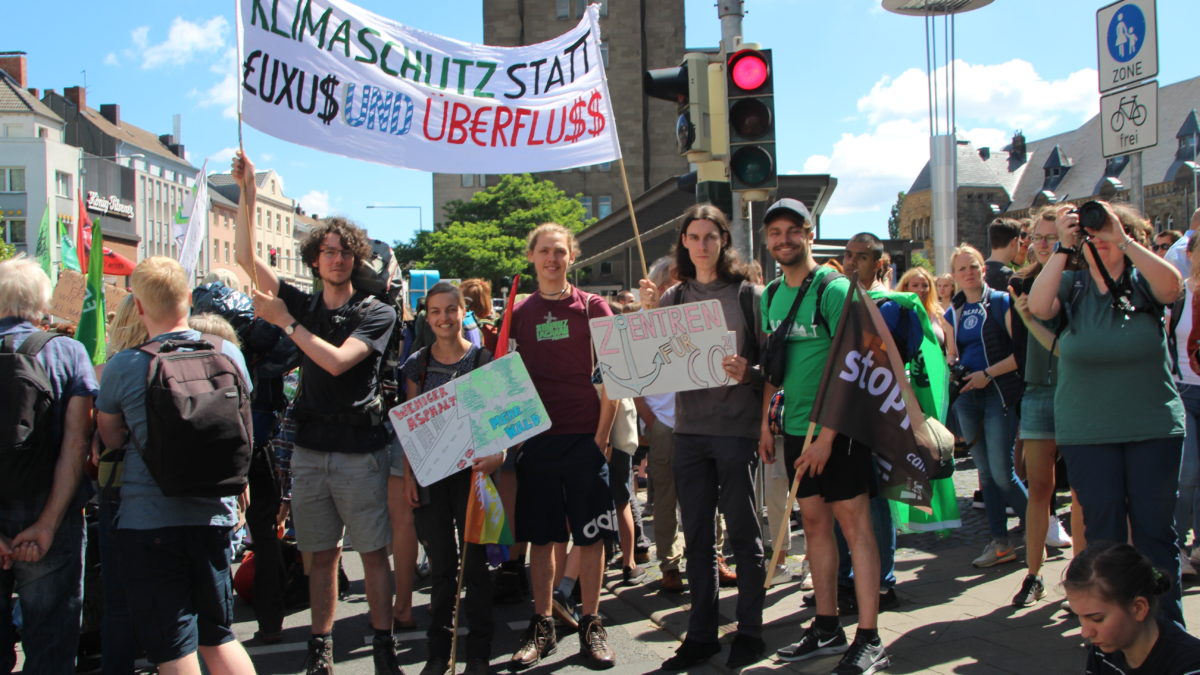 Menschen des Kaktus auf einer Demo. Sie halten ein Plakat. Auf dem Plakat steht "Klimaschutz statt Luxus und Überfluss".