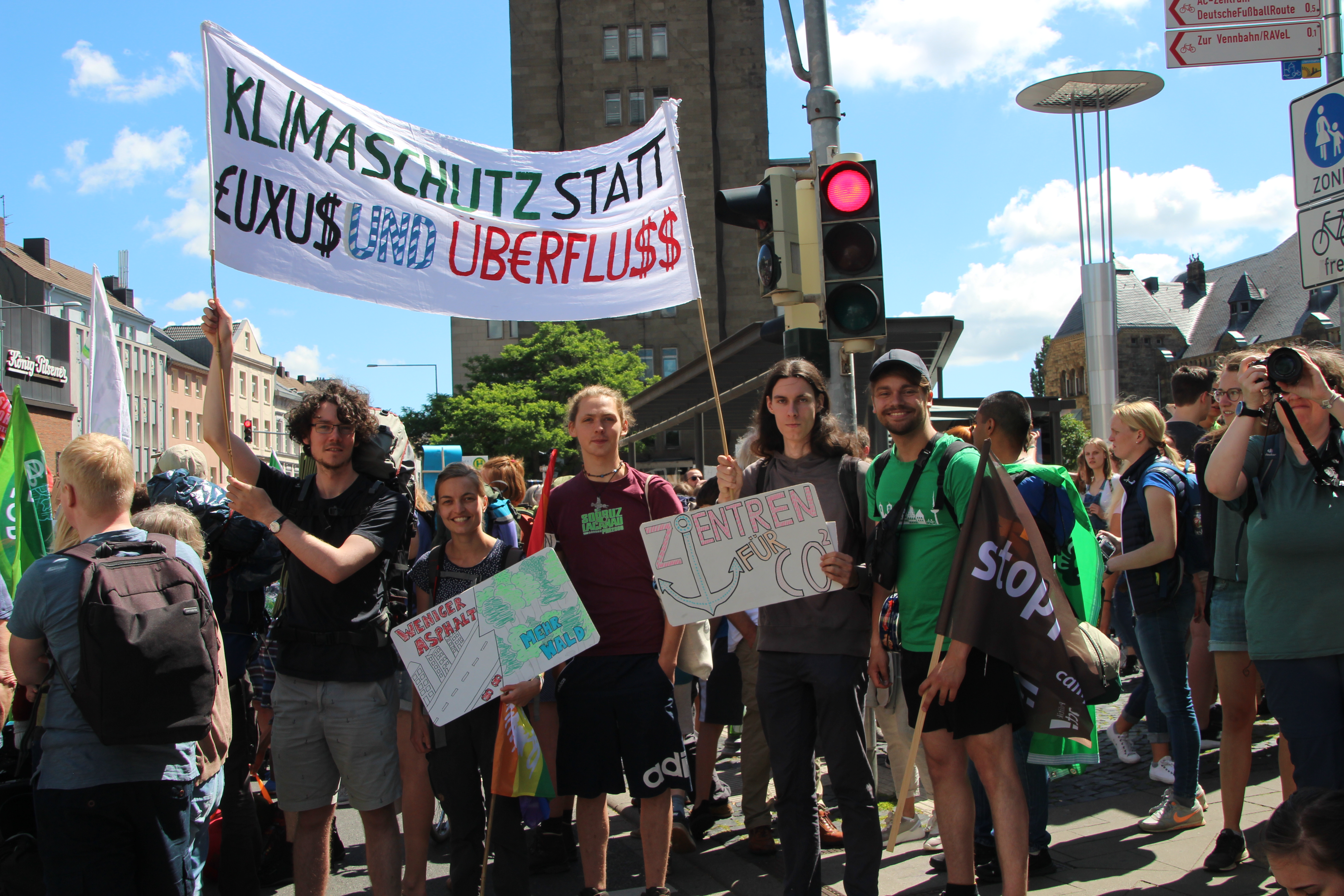 Fünf Menschen stehen in einer Menschen·menge. Sie halten ein Plakat. Auf dem Plakat steht: "Klimaschutz statt Luxus und Überfluss."