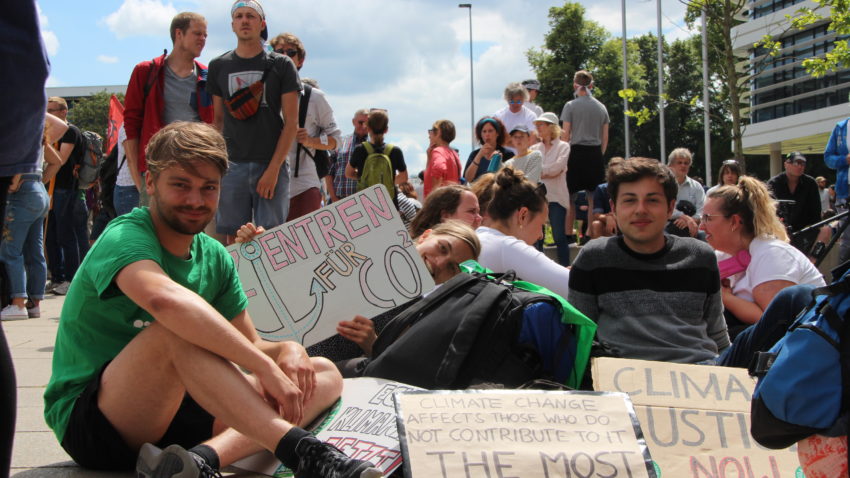 Viele Menschen sitzen auf dem Boden. Auf Plakten stehen verschiedene Sätze: "Ankerzentren für CO2." und "Climate Justice Now."