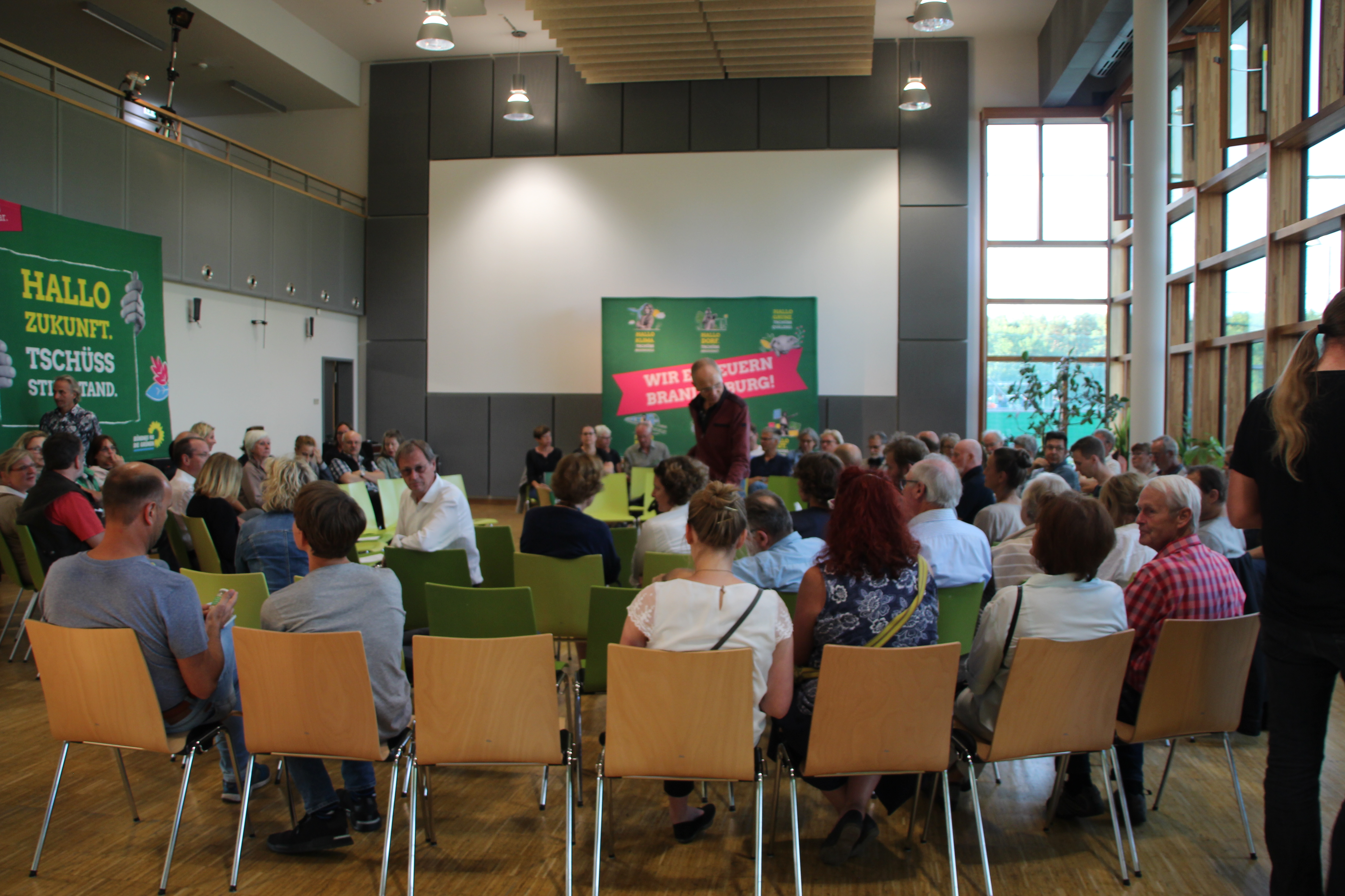 Viele Menschen sitzen auf Stühlen und reden. Sie warten auf eine Wahl·kampf·veranstaltung.