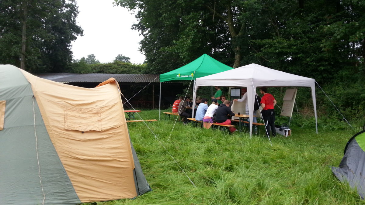 Menschen sitzen auf einer Wiese neben Zelten unter Pavillons und lauschen einem Vortrag.