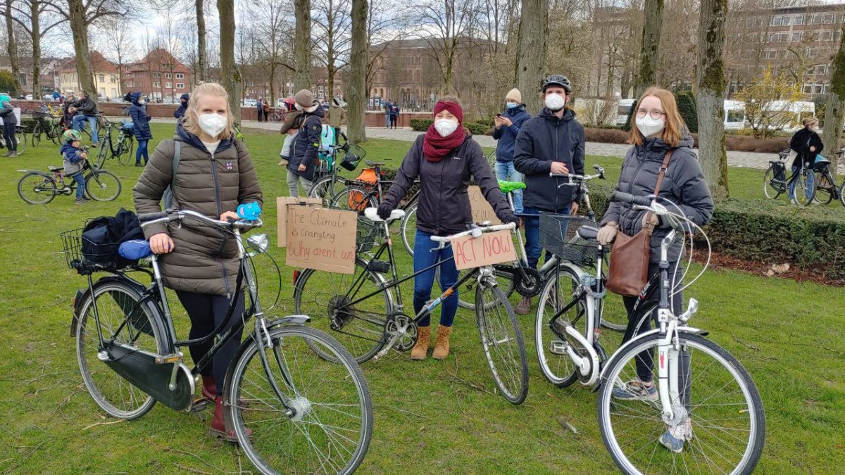 Menschen stehen mit Masken und Fahrrädern auf einer Wiese und schauen in die Kamera.