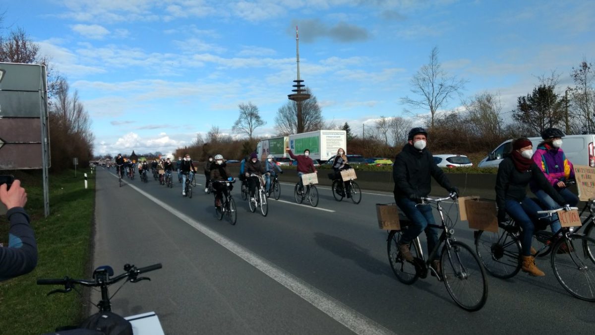 Menschen fahren mit ihren Fahrrädern über eine Bundesstraße. Manche winken. Auf der anderen Fahrbahn stehen Autos im Stau.