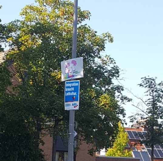 An einer Laterne hängt ein Plakat der AfD. Über dem Plakat hängt ein Plakat der Grünen Jugend Münster. Auf dem Plakat ist ein Einhorn, das einen Regenbogen nach unten auf das AfD-Plakat kotzt, zu sehen.