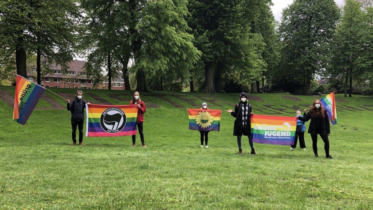 Menschen stehen mit Abstand auf einer Wiese und halten verschiedene Pride-Fahnen.