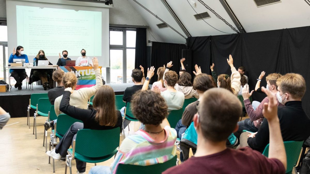 Menschen sitzen in Reihen und heben die Hand. Sie stimmen über einen Antrag ab.